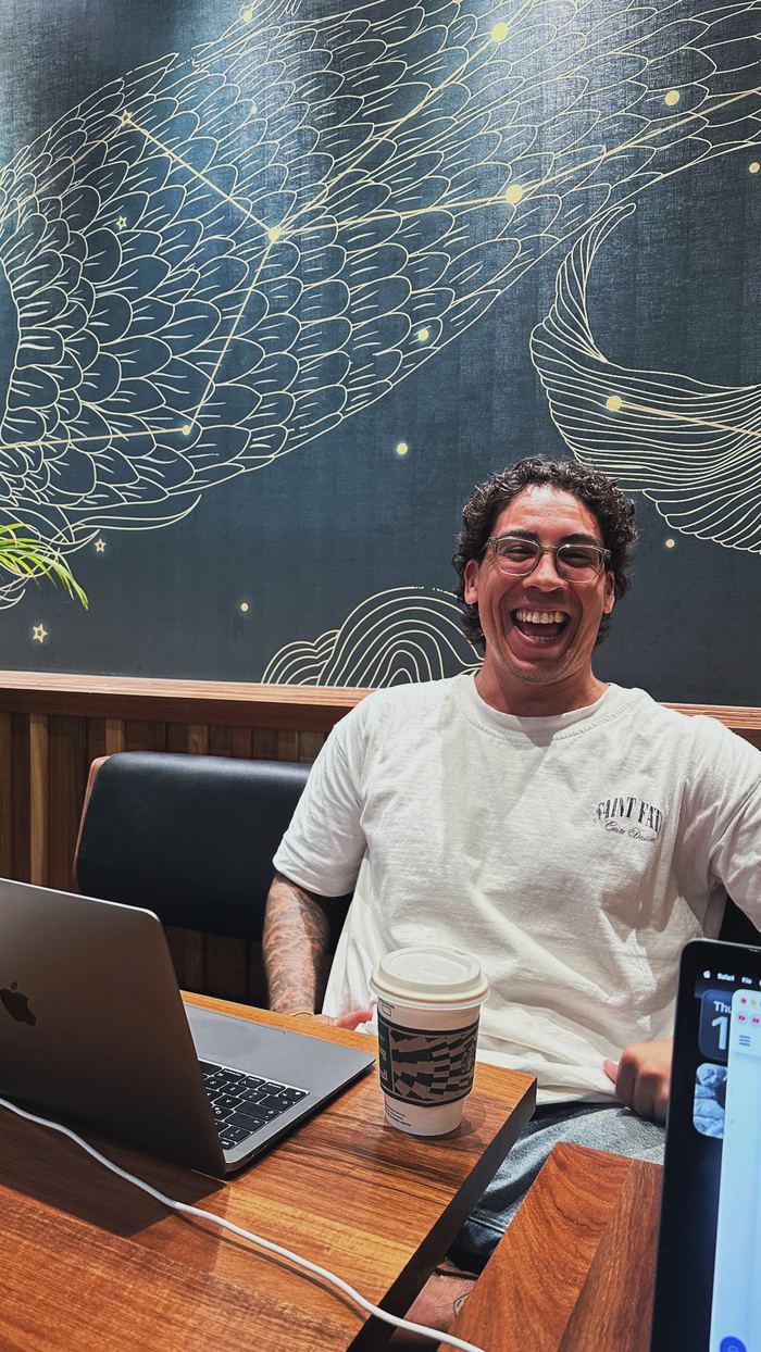 Agustín riendo frente a su laptop en una cafetería, con un café al lado y un mural de constelaciones de fondo