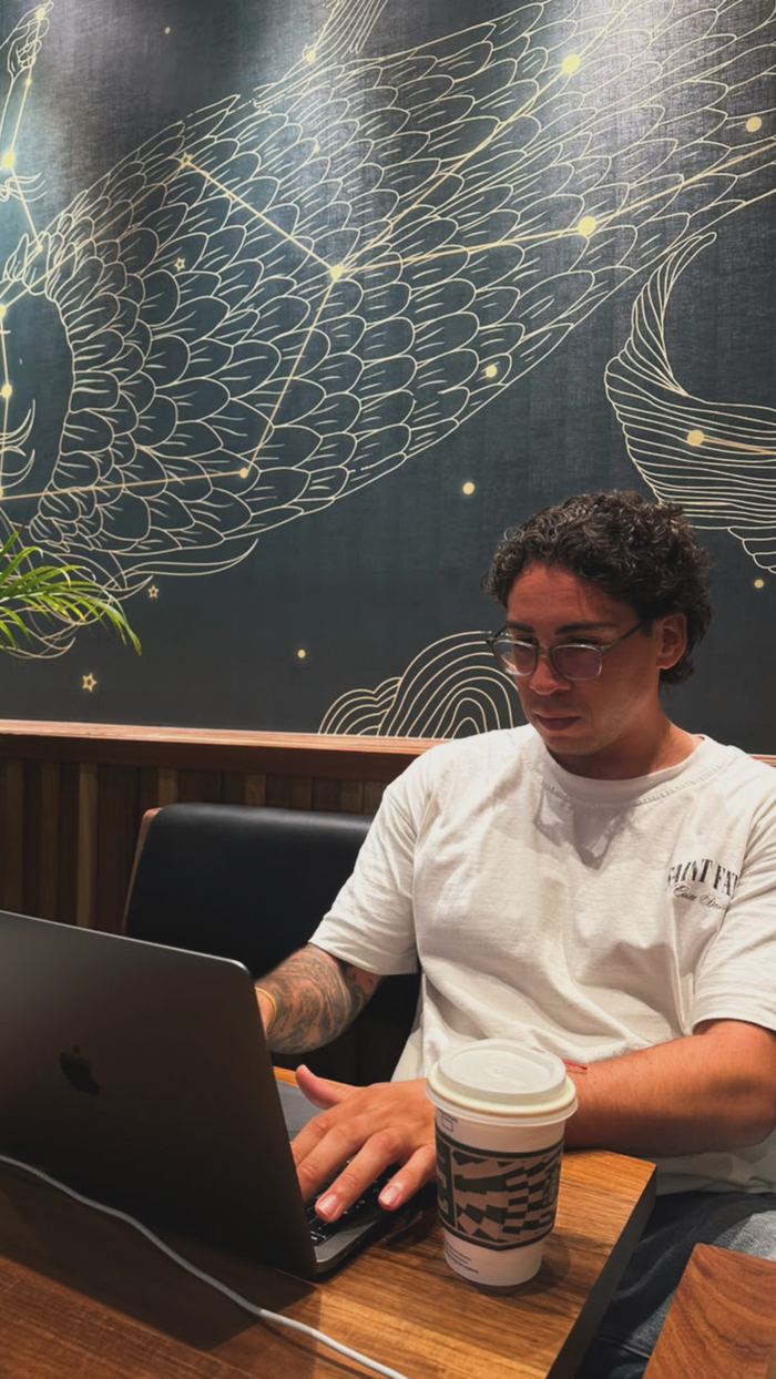 Agustín escribiendo en su laptop, concentrado, en la misma cafetería del mural de constelaciones